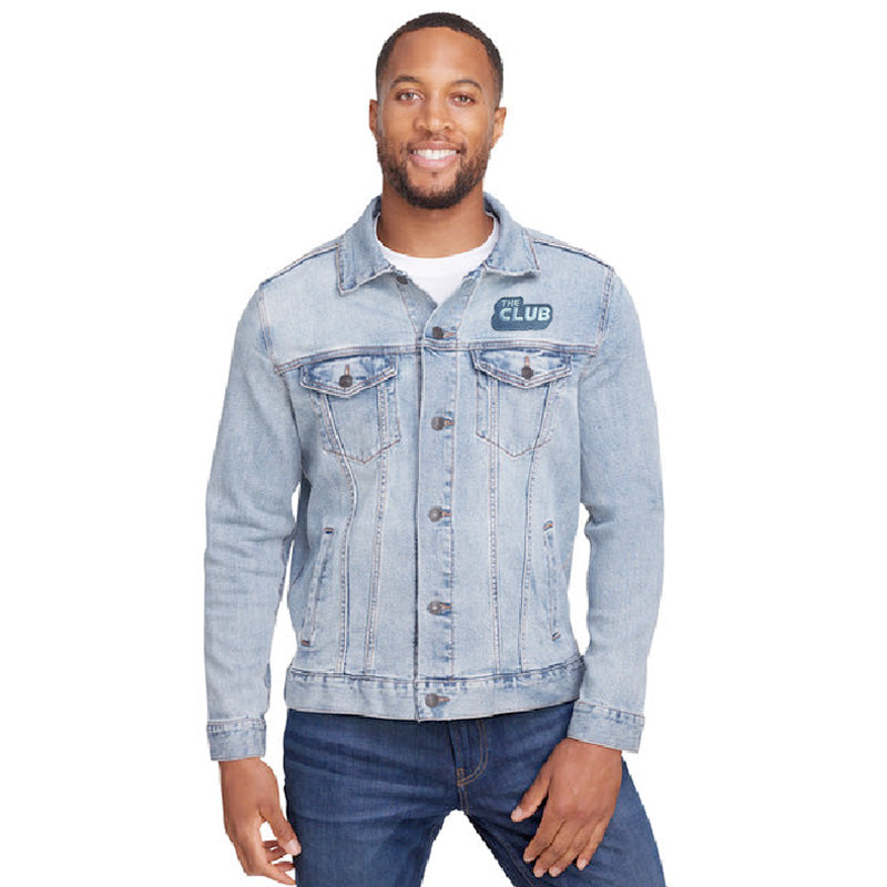 Smiling man wearing a light denim unisex jacket with “THE CLUB” patch on the left chest, buttoned up and paired with dark blue jeans.