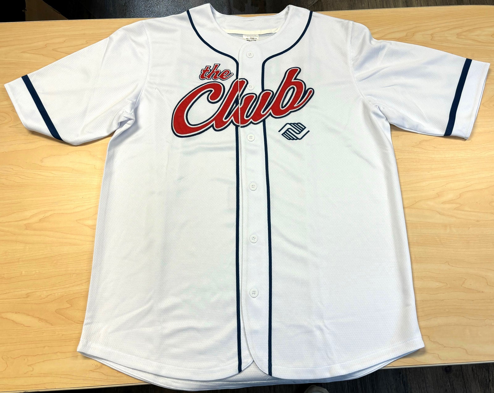 White baseball-style jersey with navy trim, featuring large red and white text reading “the Club” across the chest, and the Boys & Girls Clubs logo below.